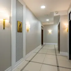 Korytarz hotelowy z jasnymi ścianami, co oznacza kolor szary, i eleganckimi lampami. Widać drzwi, windę i numer pokoju 7.