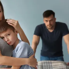 Smutna kobieta tuli syna, podczas gdy w tle stoi zmartwiony mężczyzna. Trudno jej zaakceptować dziecko partnera.