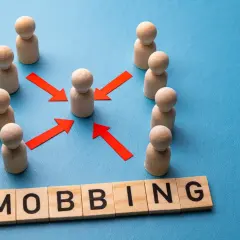 Drewniane ludziki otaczają jednego, a czerwone strzałki wskazują na niego. Słowo "MOBBING" ułożone z klocków symbolizuje nękanie w pracy.