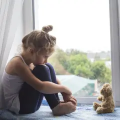 Smutna dziewczynka siedzi przy oknie, przytulając kolana. Obok niej pluszowy miś. Obraz symbolizuje wykluczenie z grupy rówieśniczej i poczucie samotności.