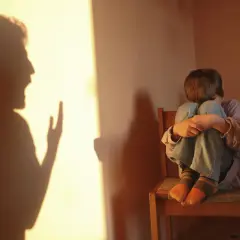 Cień krzyczącej postaci rzuca się na ścianę, podczas gdy skulone dziecko siedzi na krześle, odzwierciedlając długotrwałe skutki bicia dzieci w dorosłym życiu.