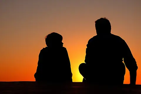 Dwie sylwetki, dziecko i dorosły, siedzą obok siebie na tle zachodzącego słońca. Obraz symbolizuje relacje z mężczyznami, nawet w obliczu braku ojca.