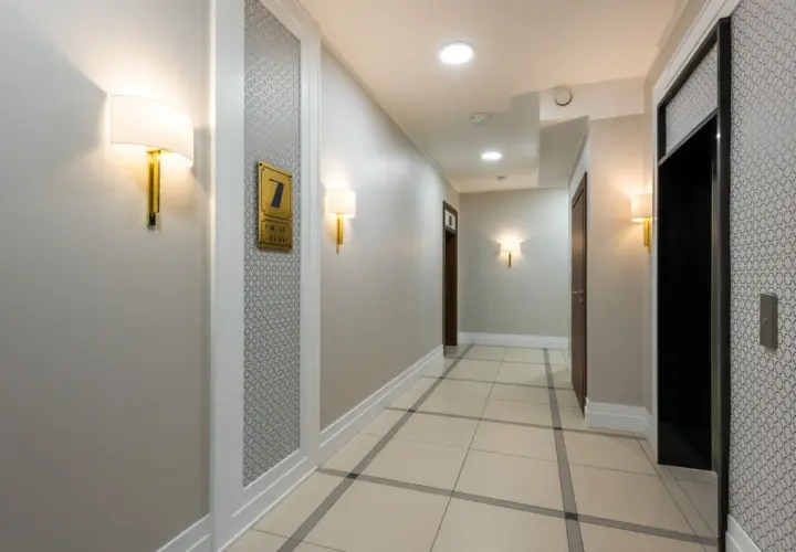 Korytarz hotelowy z jasnymi ścianami, co oznacza kolor szary, i eleganckimi lampami. Widać drzwi, windę i numer pokoju 7.