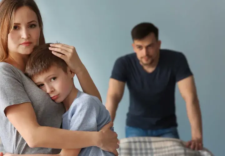 Smutna kobieta tuli syna, podczas gdy w tle stoi zmartwiony mężczyzna. Trudno jej zaakceptować dziecko partnera.
