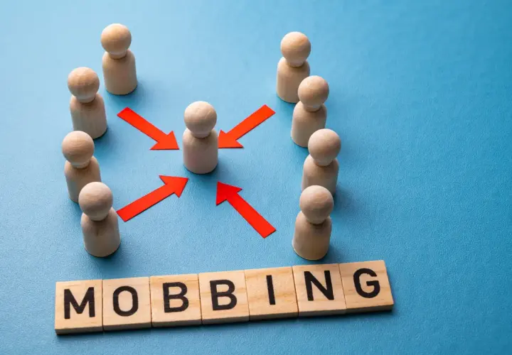 Drewniane ludziki otaczają jednego, a czerwone strzałki wskazują na niego. Słowo "MOBBING" ułożone z klocków symbolizuje nękanie w pracy.