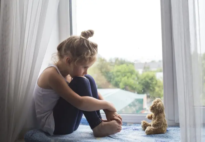 Smutna dziewczynka siedzi przy oknie, przytulając kolana. Obok niej pluszowy miś. Obraz symbolizuje wykluczenie z grupy rówieśniczej i poczucie samotności.