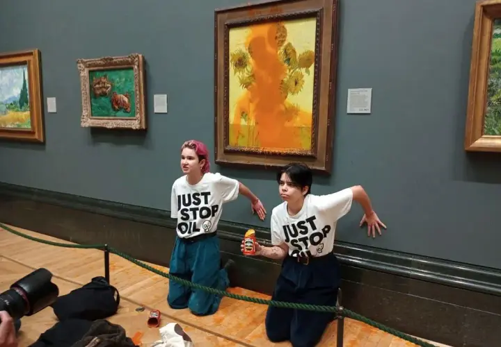 Dwie osoby w koszulkach "JUST STOP OIL" klęczą przed obrazem "Słoneczniki Van Gogha", na który wylano pomarańczową farbę.