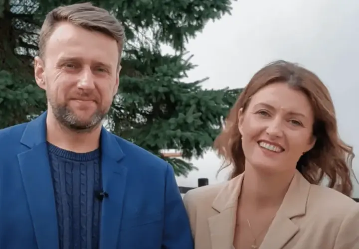 Uśmiechnięta para, on w niebieskiej marynarce, ona w beżowej. Wyglądają jak towarzystwo wzajemnej adoracji, ciesząc się wspólnym momentem.