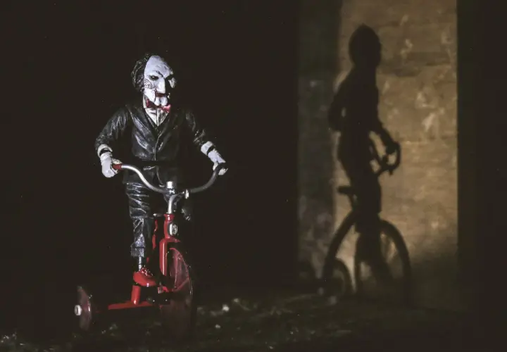 Lalka Billy z Piły na czerwonym trójkołowcu, jej cień na ścianie przypomina o najlepszych horrorach psychologicznych.
