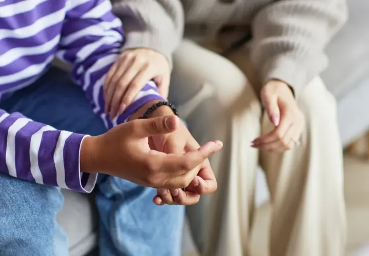 Dwie osoby siedzą blisko siebie. Jedna z nich, w bluzie w fioletowo-białe pasy, trzyma dłoń drugiej, która delikatnie ją obejmuje. To chwila, gdy można komuś zwierzac się.