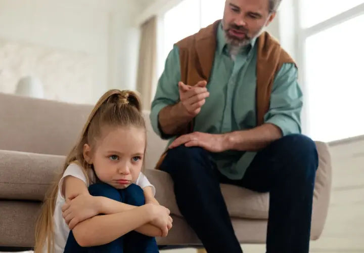 Smutna dziewczynka siedzi na podłodze, podczas gdy ojciec mówi do niej ostro, ukazując typowe daddy issues zachowania.