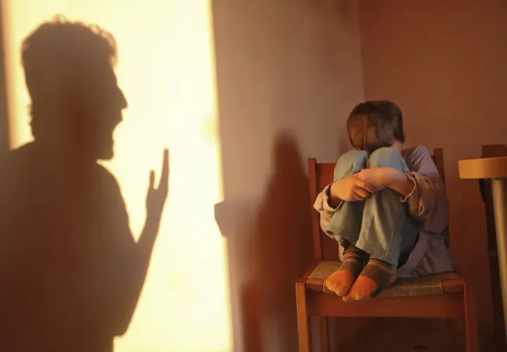 Cień krzyczącej postaci rzuca się na ścianę, podczas gdy skulone dziecko siedzi na krześle, odzwierciedlając długotrwałe skutki bicia dzieci w dorosłym życiu.