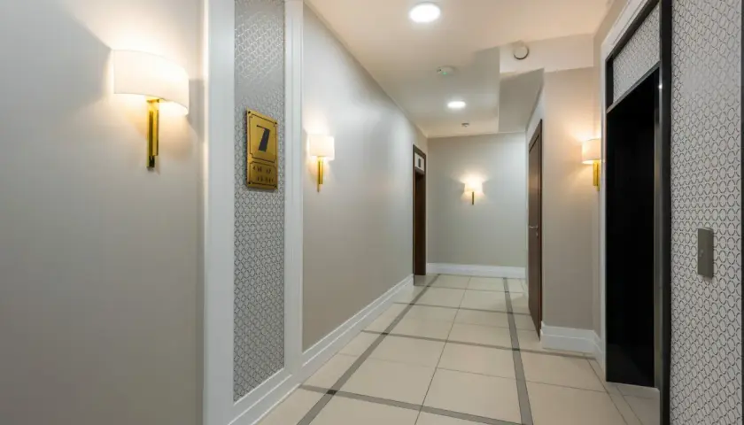 Korytarz hotelowy z jasnymi ścianami, co oznacza kolor szary, i eleganckimi lampami. Widać drzwi, windę i numer pokoju 7.