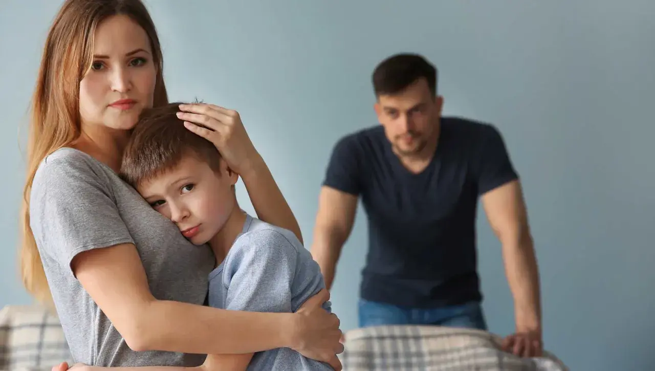 Smutna kobieta tuli syna, podczas gdy w tle stoi zmartwiony mężczyzna. Trudno jej zaakceptować dziecko partnera.