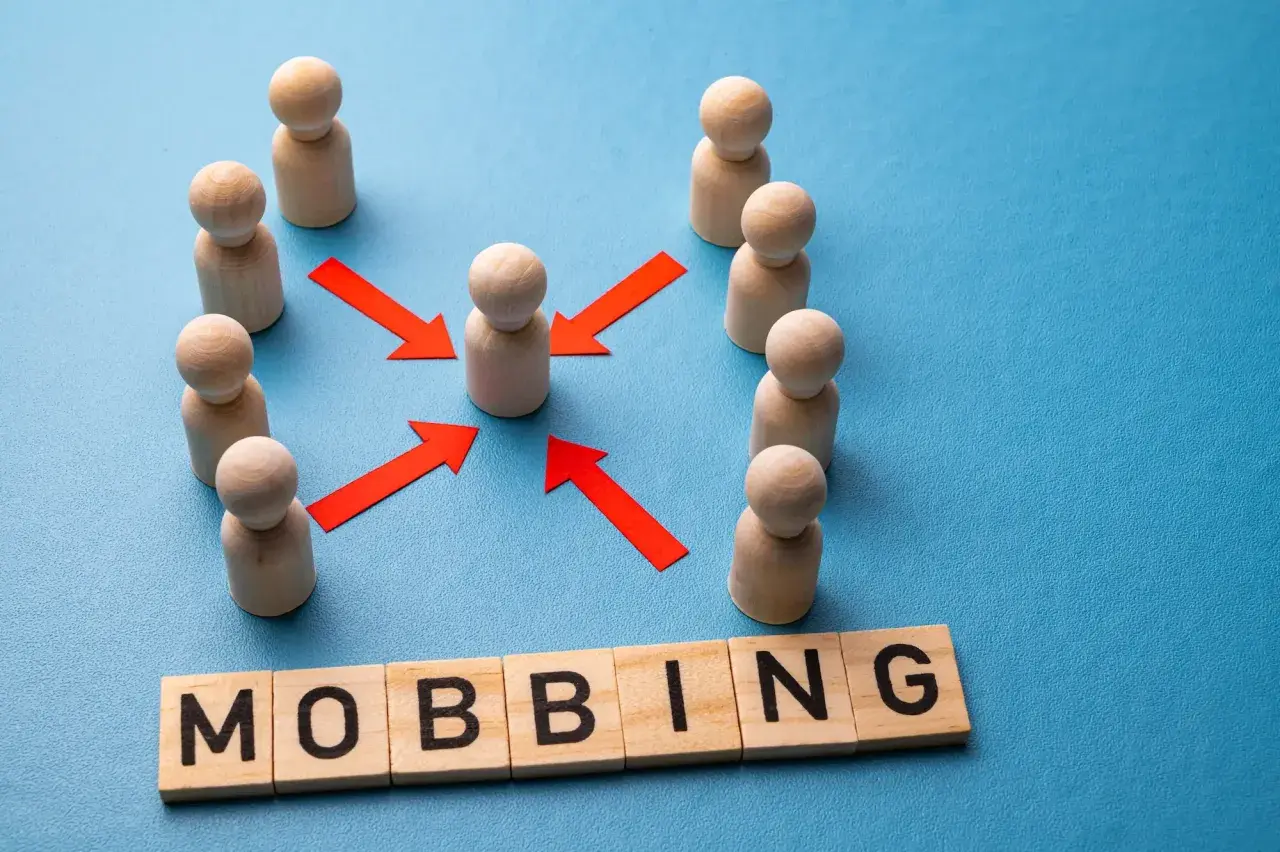 Drewniane ludziki otaczają jednego, a czerwone strzałki wskazują na niego. Słowo "MOBBING" ułożone z klocków symbolizuje nękanie w pracy.