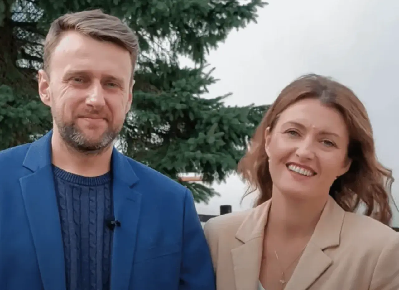 Uśmiechnięta para, on w niebieskiej marynarce, ona w beżowej. Wyglądają jak towarzystwo wzajemnej adoracji, ciesząc się wspólnym momentem.