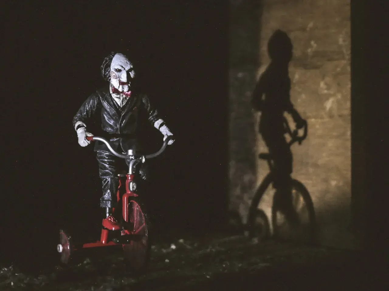 Lalka Billy z Piły na czerwonym trójkołowcu, jej cień na ścianie przypomina o najlepszych horrorach psychologicznych.