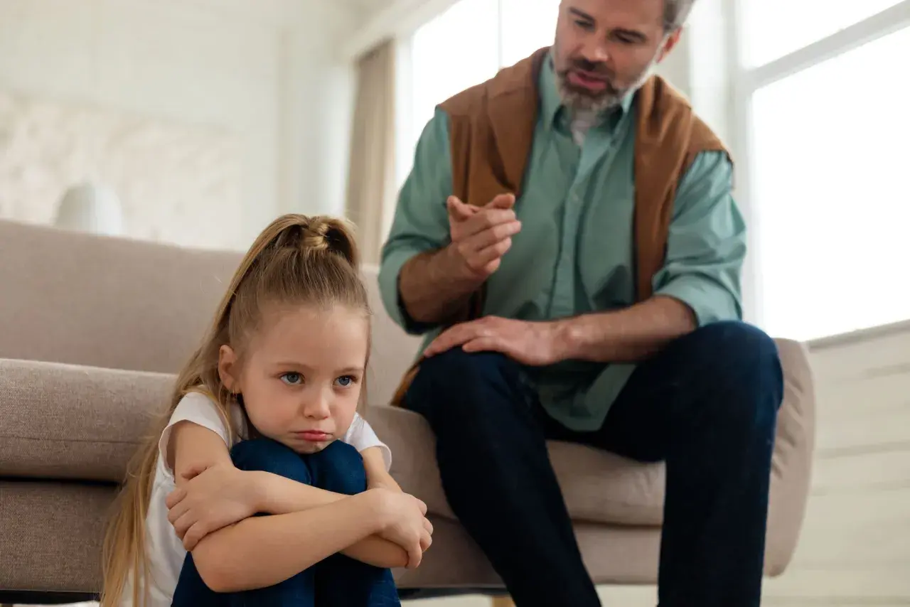 Smutna dziewczynka siedzi na podłodze, podczas gdy ojciec mówi do niej ostro, ukazując typowe daddy issues zachowania.