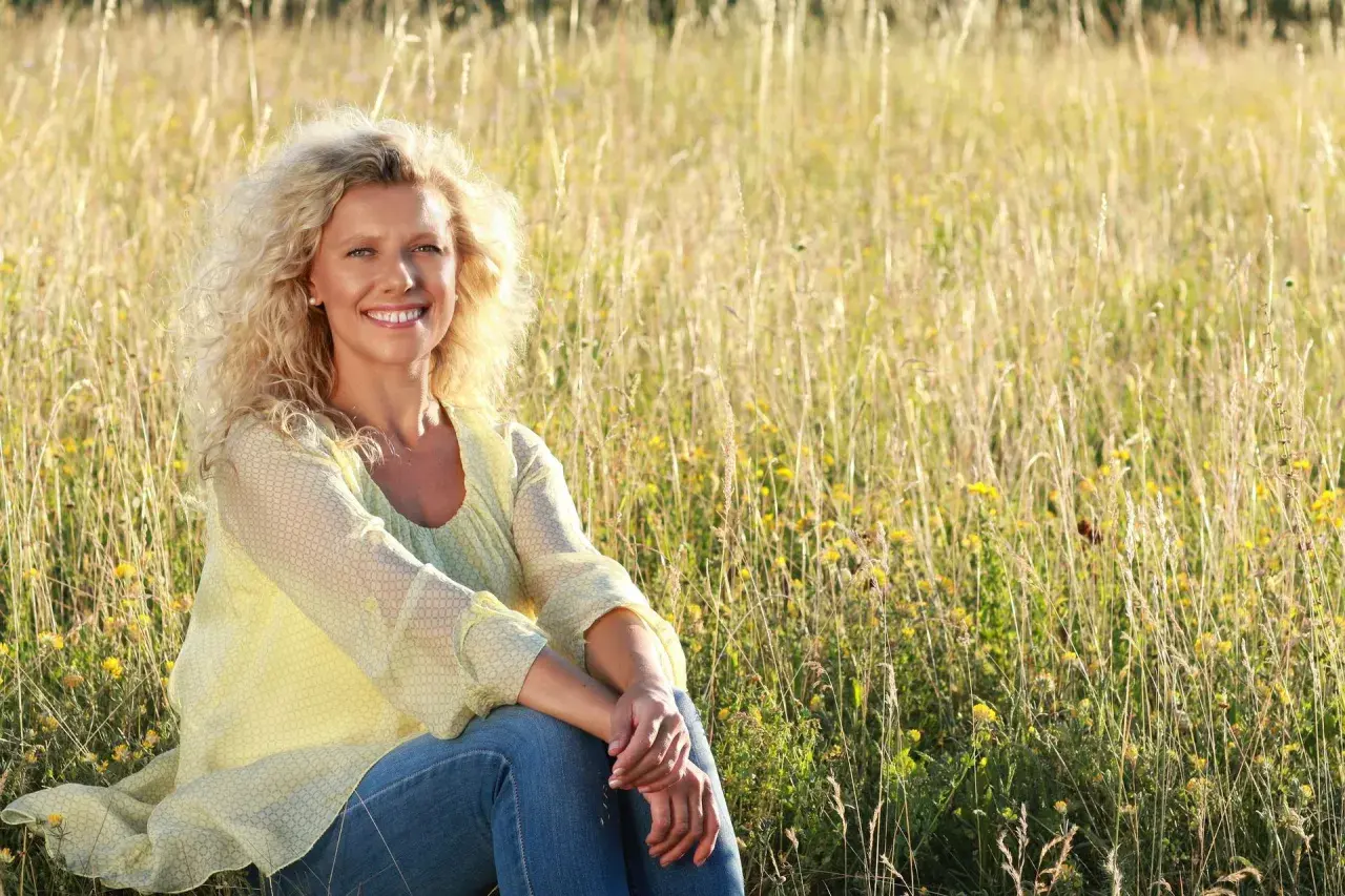 Uśmiechnięta kobieta po 50. siedzi na łące pełnej traw i kwiatów. Jestem samotna po 50. i cieszę się chwilą spokoju.