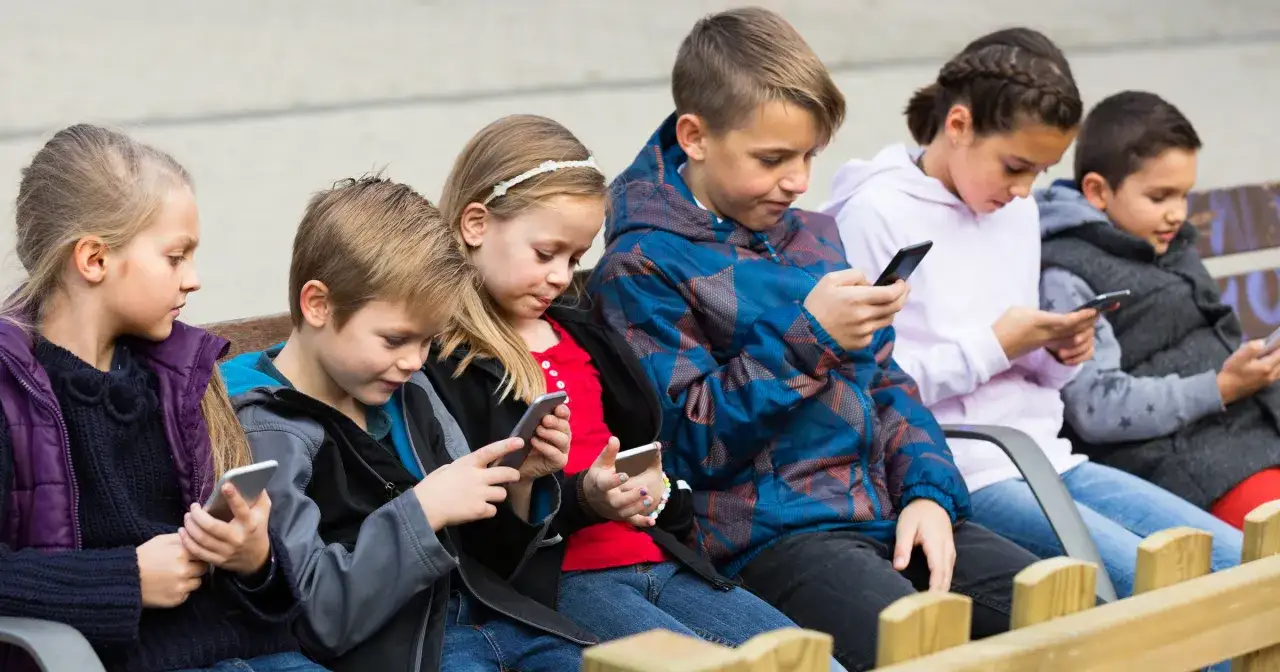 Dzieci siedzą na ławce, każde wpatrzone w swój smartfon. Czy to efekt wertera, co to znaczy dla ich relacji?