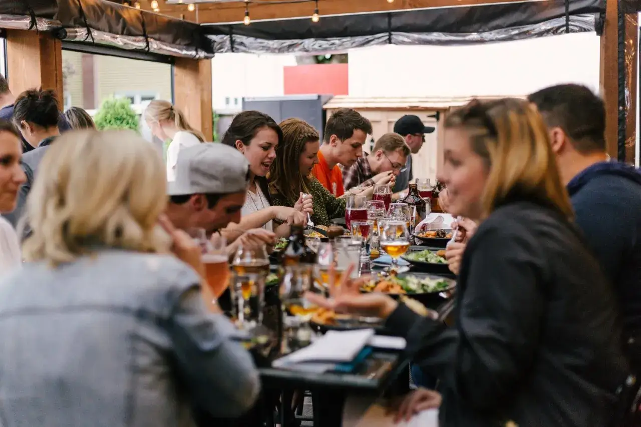 Ludzie jedzą i piją przy długim stole, tworząc żywy efekt cocktail party.