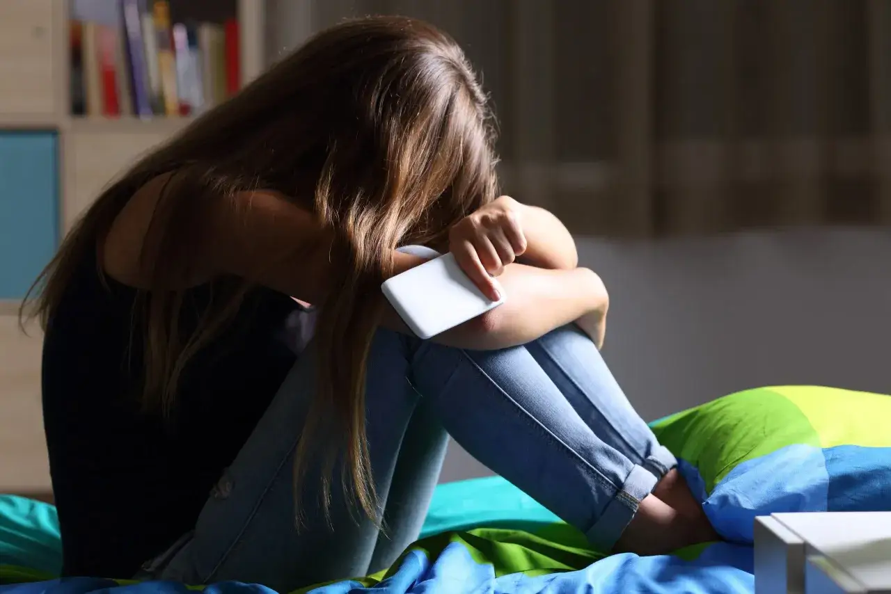 Dziewczyna siedzi skulona na łóżku, trzymając telefon. Wokół niej wszystko się układa, tylko nie jej.