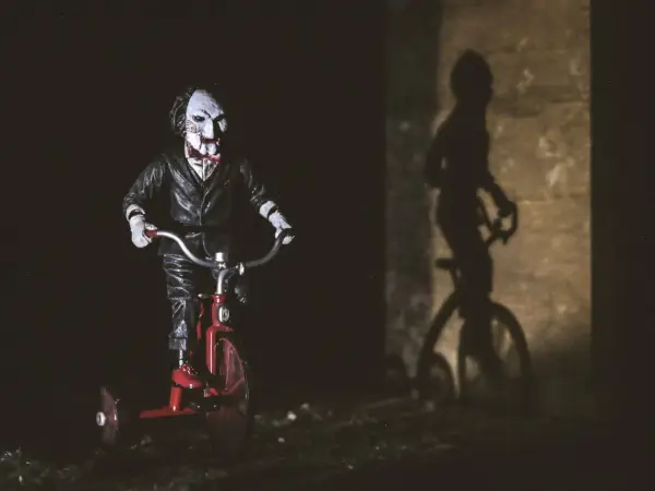 Lalka Billy z Piły na czerwonym trójkołowcu, jej cień na ścianie przypomina o najlepszych horrorach psychologicznych.